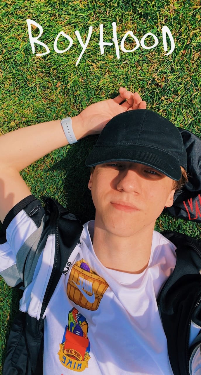 Selfie de moi dans l'herbe, avec un tee shirt blanc, une veste de sport à manche courte noir, grise et blanche. J'ai une casquette noir, le soleil qui aurait dû m'éblouir sans celle-ci, et la main droite derrière la tête, allongée sur du beau gazon vert.